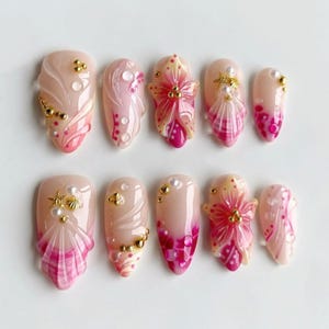May include: A set of ten artificial nails with a light pink base. Each nail features intricate designs, including seashell and floral embellishments, with gold accents and pearl details. The nails showcase a gradient of pink and white.