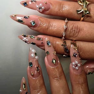 May include: Long almond-shaped nails with a sheer nude base coat, decorated with various multi-colored rhinestones, small white pearls, and gold metal studs. The fingers are adorned with a gold bow ring and a thin band with clear stones.
