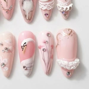 May include: A set of almond-shaped press-on nails in shades of pale pink. The designs include hand-painted white swans, delicate floral stems, and white lace-like textures. Each nail is embellished with small, clear, reflective rhinestones.