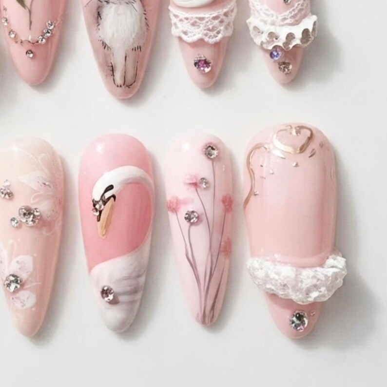 May include: A set of almond-shaped press-on nails in shades of pale pink. The designs include hand-painted white swans, delicate floral stems, and white lace-like textures. Each nail is embellished with small, clear, reflective rhinestones.