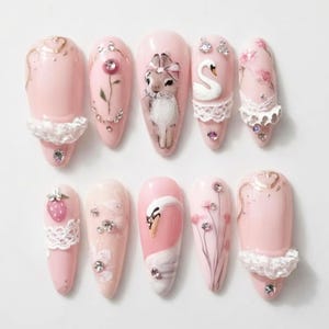 May include: A set of ten almond-shaped press-on nails in shades of soft pink. The designs feature hand-painted rabbits, white swans, floral motifs, and strawberries, accented with white lace trim, clear rhinestones, and metallic gold line art.
