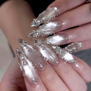 May include: Long, almond-shaped nails featuring a shimmering, pearlescent light pink polish. Each nail is intricately decorated with clusters of clear, multifaceted rhinestones and small silver beads concentrated near the tips and cuticle areas.