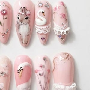 May include: A set of eight almond-shaped press-on nails in a soft pink base color. The designs feature hand-painted illustrations of a rabbit with a bow, white swans, and floral motifs. Embellishments include white lace trim and small clear rhinestones.