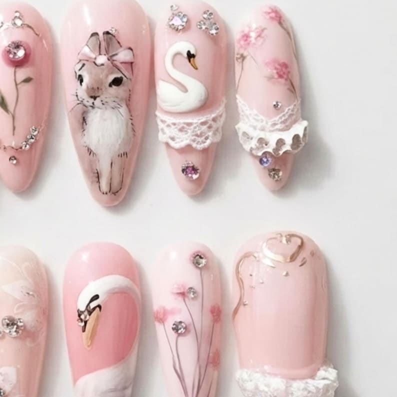 May include: A set of eight almond-shaped press-on nails in a soft pink base color. The designs feature hand-painted illustrations of a rabbit with a bow, white swans, and floral motifs. Embellishments include white lace trim and small clear rhinestones.