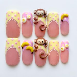 May include: A set of ten press-on nails featuring a French manicure style with pink bases and yellow tips adorned with pink polka dots and white scalloped edges. The design includes 3D embellishments of brown monkeys, yellow bananas, and sliced fruit.