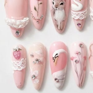 May include: A set of almond-shaped press-on nails in shades of soft pink. The designs feature white lace trim, clear rhinestones, hand-painted illustrations of a rabbit, a swan, a strawberry, and delicate floral stems on a smooth glossy finish.
