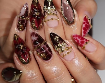 May include: Stiletto-shaped artificial nails featuring a mix of deep red, dark brown, and nude base colors. The designs include 3D red floral accents, gold metallic cross charms, intricate gold bead chains, and glossy dark red crocodile-like textures.