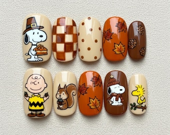 May include: A set of ten press-on nails with fall-themed designs. The nails feature cartoon characters Snoopy and Charlie Brown, along with autumn leaves, squirrels, and checkerboard patterns. Colors include beige, brown, orange, and yellow.