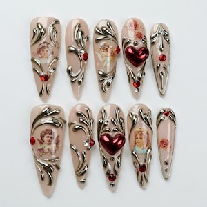 May include: A set of ten almond-shaped press-on nails in a nude base color. The nails feature raised silver metallic scrollwork, red heart-shaped charms, small red and clear rhinestones, and printed images of cherubs and red roses on select nails.