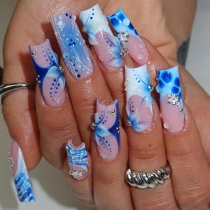 May include: Long square-shaped artificial nails featuring a French manicure base with intricate blue and white floral patterns, swirling lines, and small silver metallic studs. The design includes raised clear droplet accents and small silver flower charms.