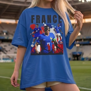 May include: Blue t-shirt featuring the word "FRANCE" in black block letters above a graphic of French soccer players in action. The shirt has a crew neck and short sleeves. The background is a soccer field.