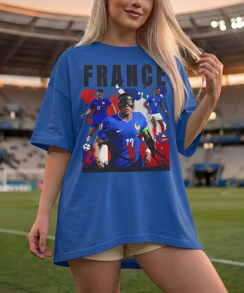 May include: Blue t-shirt featuring the word "FRANCE" in black block letters above a graphic of French soccer players in action. The shirt has a crew neck and short sleeves. The background is a soccer field.