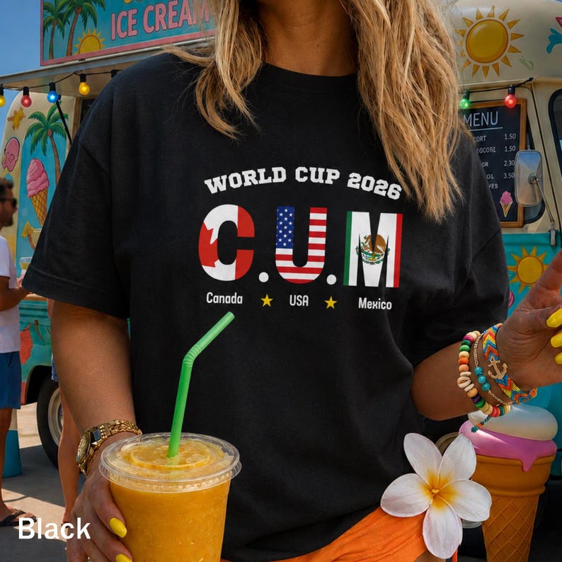 Op de afbeelding: Een zwart t-shirt met de tekst "WORLD CUP 2026" boven de letters "C.U.M." die zijn gevuld met de vlaggen van Canada, de VS en Mexico. Onder de letters staan de woorden "Canada", "USA" en "Mexico" gedrukt met gele sterren.