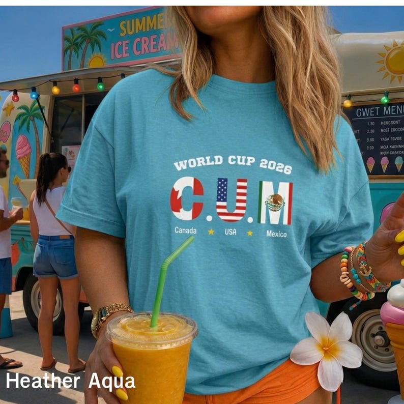 Op de afbeelding: Een lichtblauw t-shirt met de tekst WORLD CUP 2026 boven de letters C, U en M, die zijn gevuld met de vlaggen van Canada, de VS en Mexico. Onder de letters staan de woorden Canada, USA en Mexico gedrukt met gele sterren.
