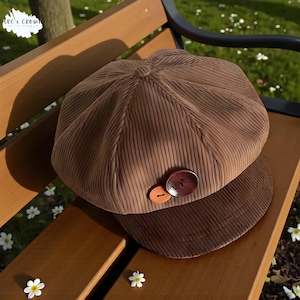 Könnte beinhalten: Braune Cord-Newsboy-Cap mit runder Krone und kurzem, steifem Schirm. Die Cap hat einen dekorativen Holzknopf und einen kleineren orangefarbenen Knopf. Die Mütze liegt auf einer Holzbank in einer Parkumgebung.