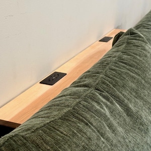 May include: A light-colored wooden headboard shelf featuring two black rectangular power outlet inserts, positioned directly behind a textured, olive green upholstered headboard with vertical stitched detailing.