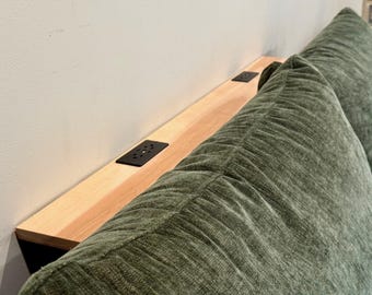 May include: A light-colored wooden headboard shelf featuring two black rectangular power outlet inserts, positioned directly behind a textured, olive green upholstered headboard with vertical stitched detailing.