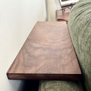 May include: A rectangular wooden armrest tray with a dark brown finish and visible natural wood grain patterns. The flat surface is positioned horizontally across the arm of a textured olive green upholstered sofa, creating a stable platform.