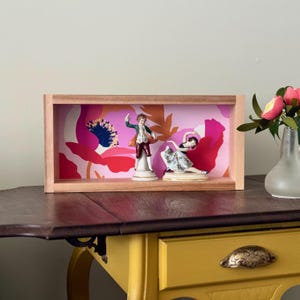 May include: A wooden framed display box featuring two porcelain figurines of dancers against a floral backdrop. The figurines are dressed in period clothing. The background is a pink and red floral pattern.