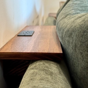 May include: A wooden sofa arm table with a dark stain. The table has a square, dark-colored accent piece on the surface. The table is positioned next to a light-colored wall and a green sofa.