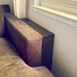 May include: A dark brown wooden console table positioned in the narrow space between a brown upholstered sofa arm and a white wall. The rectangular table features a smooth, flat top surface and a vertical side panel that fits flush against the sofa.