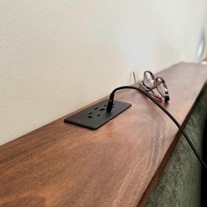 May include: A dark brown wooden shelf with a black power outlet and a charging cable. A pair of eyeglasses rests on the shelf. The shelf is mounted on a wall next to a green sofa.