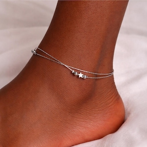 May include: A silver-coloured, layered anklet featuring a star charm and small bead accents. The anklet is worn on the ankle. The jewellery is delicate and minimalist.