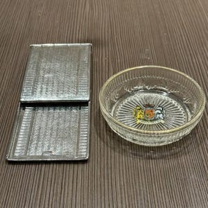 Pode incluir: Um porta-cigarros prateado com um padrão texturizado e uma pequena tigela de vidro com borda dourada e um brasão. A tigela tem um padrão radial. Em uma superfície de madeira.