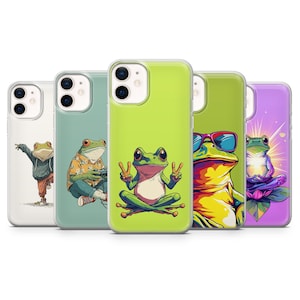 May include: A collection of five phone cases featuring colorful frog illustrations. Each case showcases a different frog design, including one with sunglasses, one in a hoodie, and one with peace signs. The cases are in various colors, including green, purple, and white.