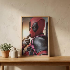 Op de afbeelding: Een ingelijste muurkunstprint met een personage dat een rood en zwart masker met witte oogvlekken draagt, met een gehandschoende vinger voor de lippen in een stiltegebaar. Het personage draagt een zwart tactisch pak met rode accenten en zwaarden.