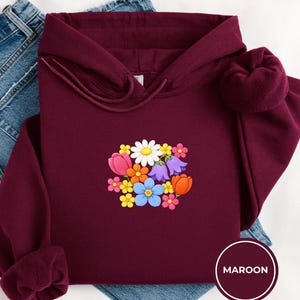 Pode incluir: Moletom bordô com um design floral com margaridas, tulipas e outras flores coloridas. O moletom com capuz é exibido em cima de um par de jeans azuis. A palavra "MAROON" está em um círculo no canto inferior direito.