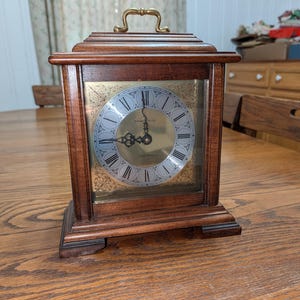 Puede incluir: Un reloj de sobremesa de madera con acabado marrón oscuro, que presenta una esfera dorada con números romanos negros y detalles de filigrana en las esquinas. El reloj tiene un asa de color latón en la parte superior y manecillas negras.
