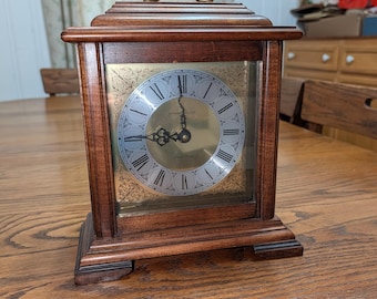 Könnte beinhalten: Eine hölzerne Kaminuhr mit dunkelbraunem Finish, die ein goldfarbenes Zifferblatt mit schwarzen römischen Ziffern und verzierten Filigran-Eckakzenten aufweist. Die Uhr hat einen messingfarbenen Griff oben und schwarze Zeiger aus Metall.