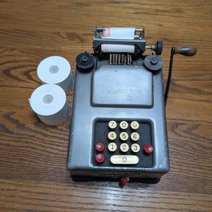 May include: A vintage Remington adding machine in a grey metal casing with a manual hand crank, a numeric keypad featuring white and red buttons, and a paper roll feed mechanism. Two additional white paper rolls are positioned to the left side.