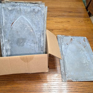 May include: A collection of rectangular metal panels with a weathered gray finish and a raised pointed arch design. Several panels are stacked inside a brown cardboard box, while one matching panel lies flat on the surface next to the box.