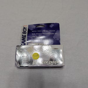 May include: Two packages of Game Boy Printer paper. The top package is dark blue with white text that reads "REFILL YOUR GAME BOY PRINTER WITH OFFICIAL NINTENDO GAME BOY PRINTER." The bottom package is silver with the text "printer paper."
