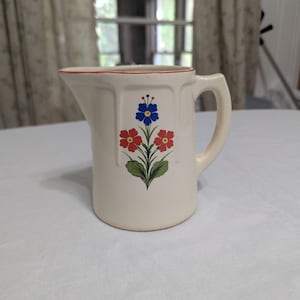 May include: Cream ceramic pitcher with a red rim and a floral design. The design features three flowers in red and blue with green leaves. The pitcher has a handle and a spout.