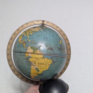 May include: A vintage globe with a blue and yellow map, featuring the continents and oceans. The globe is held by a black base and has a brass-colored meridian ring with degree markings. The word "ATLANTIC" is visible.