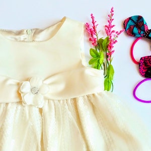 Puede incluir: Un vestido blanco sin mangas con un corpiño de satén y una falda de encaje. El vestido tiene un adorno de flores en el corpiño. También se muestran tres lazos para el pelo con lazos de colores estampados.