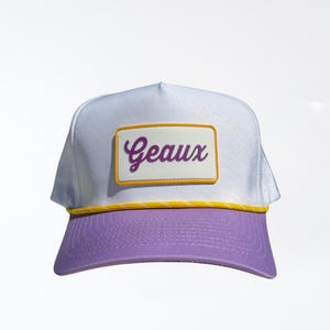 May include: White and purple trucker hat with the word "Geaux" in a purple script on a rectangular patch. The hat has a yellow trim and a purple brim.