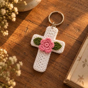 May include: A white crocheted cross keychain featuring a central pink flower with five petals and two small green leaves on either side. The cross is attached to a circular silver-toned metal split ring for keys, showcasing detailed yarn texture.
