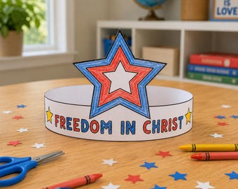 May include: A paper crown featuring a large star colored with red and blue pencil, with the text "FREEDOM IN CHRIST" printed in black on the band. The crown is surrounded by scattered paper stars, crayons, and blue scissors on a wooden surface.
