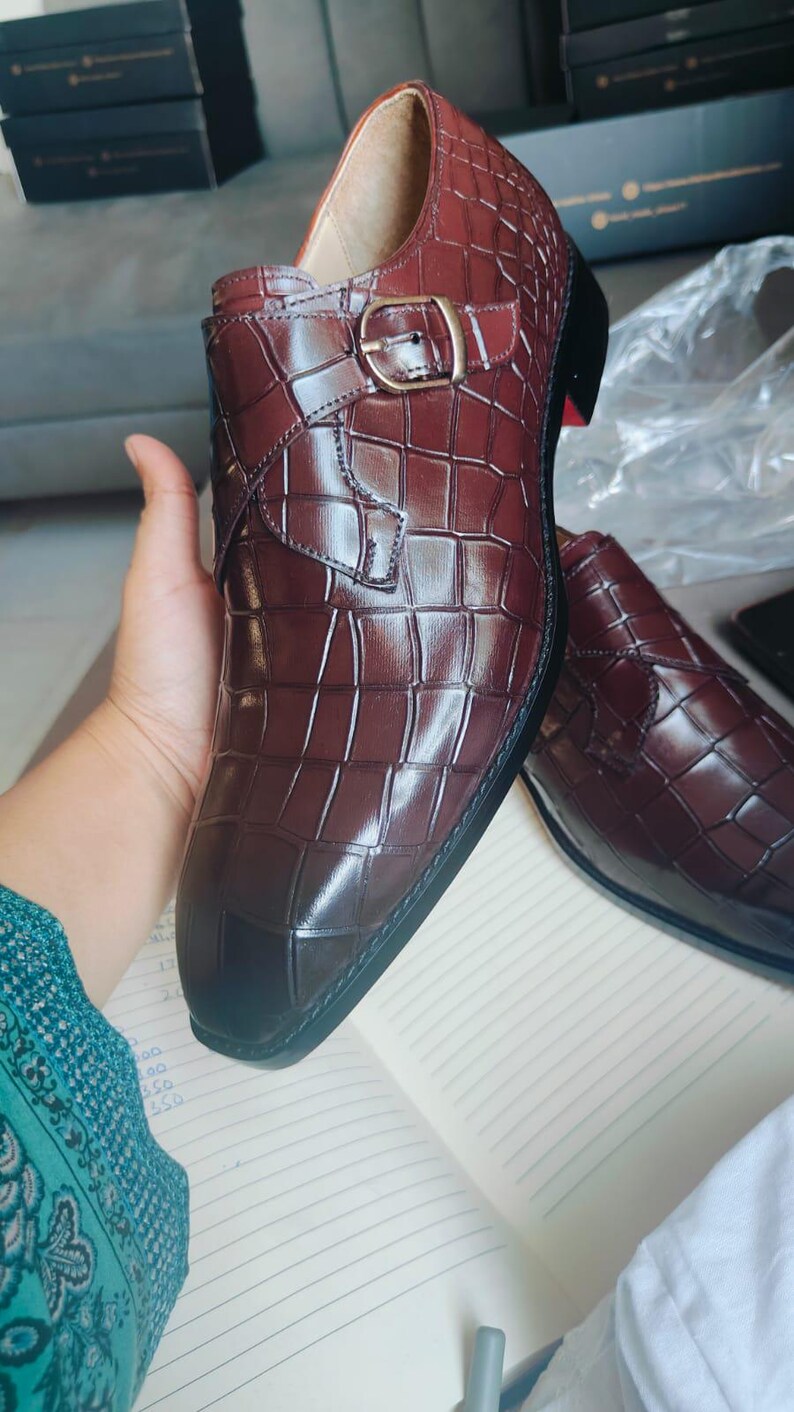 K&ouml;nnte beinhalten: Burgunderfarbene Lederschuhe mit Krokodilmuster. Die Schuhe haben einen Schnallenverschluss und eine schwarze Sohle. Die Innenseite ist in einem helleren Braunton gehalten. Die Schuhe werden auf einem Notizbuch pr&auml;sentiert.