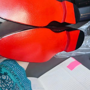 K&ouml;nnte beinhalten: Paar Herrenschuhe mit dunkelbraunem Obermaterial und leuchtend roten Sohlen. Die Sohlen sind gl&auml;nzend rot und kontrastieren mit der schwarzen Ferse. Die Schuhe werden vor einem neutralen Hintergrund pr&auml;sentiert.