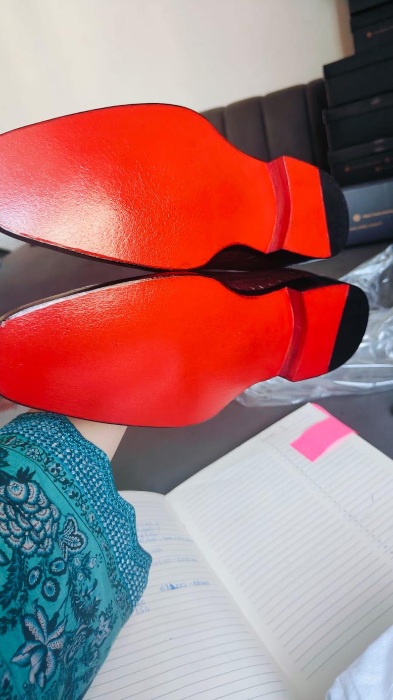 K&ouml;nnte beinhalten: Paar Herrenschuhe mit dunkelbraunem Obermaterial und leuchtend roten Sohlen. Die Sohlen sind gl&auml;nzend rot und kontrastieren mit der schwarzen Ferse. Die Schuhe werden vor einem neutralen Hintergrund pr&auml;sentiert.