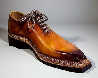 May include: A single brown leather dress shoe with a square toe and decorative stitching around the sole. The shoe has a gradient color, transitioning from light brown to a darker shade. The laces are a light brown and white.