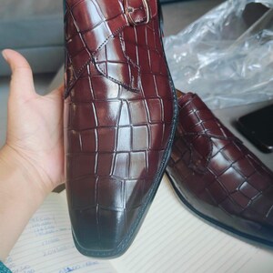 K&ouml;nnte beinhalten: Burgunderfarbene Lederschuhe mit Krokodilleder-Optik. Die Schuhe haben einen Schnallenverschluss und eine polierte Oberfl&auml;che. Das Bild zeigt die Schuhe auf einem Notizbuch mit handschriftlichen Notizen.