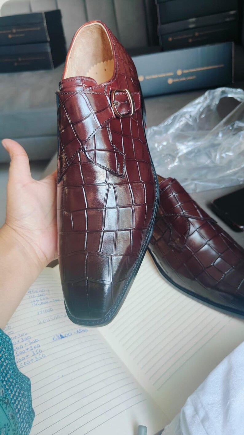 K&ouml;nnte beinhalten: Burgunderfarbene Lederschuhe mit Krokodilleder-Optik. Die Schuhe haben einen Schnallenverschluss und eine polierte Oberfl&auml;che. Das Bild zeigt die Schuhe auf einem Notizbuch mit handschriftlichen Notizen.