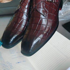 K&ouml;nnte beinhalten: Burgunderfarbene Lederschuhe mit Krokodilmuster. Die Schuhe haben einen Schnallenverschluss und eine schwarze Sohle mit rotem Akzent. Die Schuhe werden auf einem Notizbuch pr&auml;sentiert.