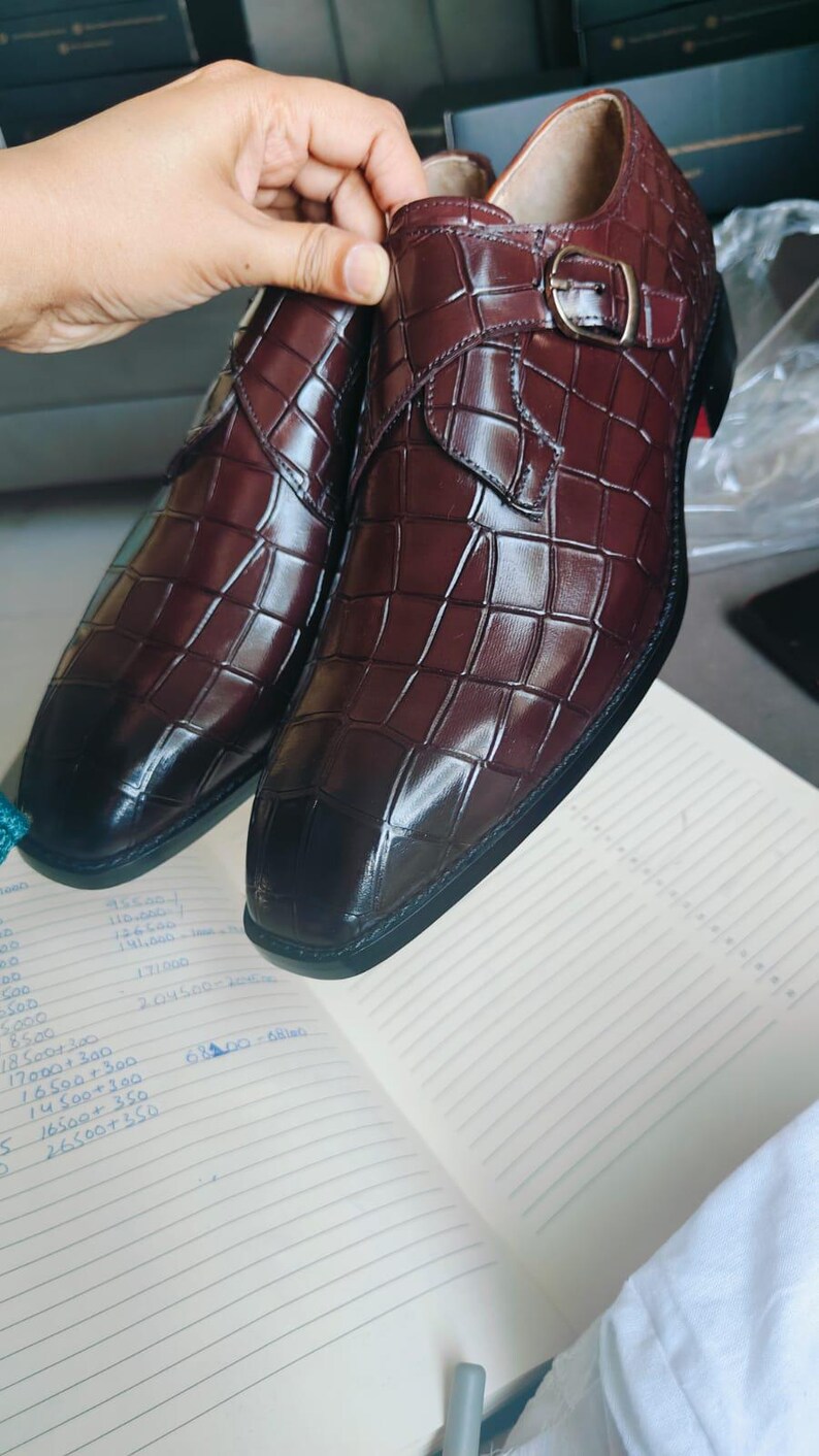 K&ouml;nnte beinhalten: Burgunderfarbene Lederschuhe mit Krokodilmuster. Die Schuhe haben einen Schnallenverschluss und eine schwarze Sohle mit rotem Akzent. Die Schuhe werden auf einem Notizbuch pr&auml;sentiert.